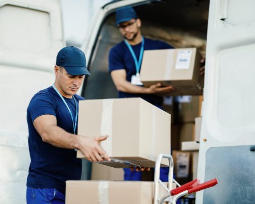 Young courier and his colleague unloading cardboard boxes from delivery van.